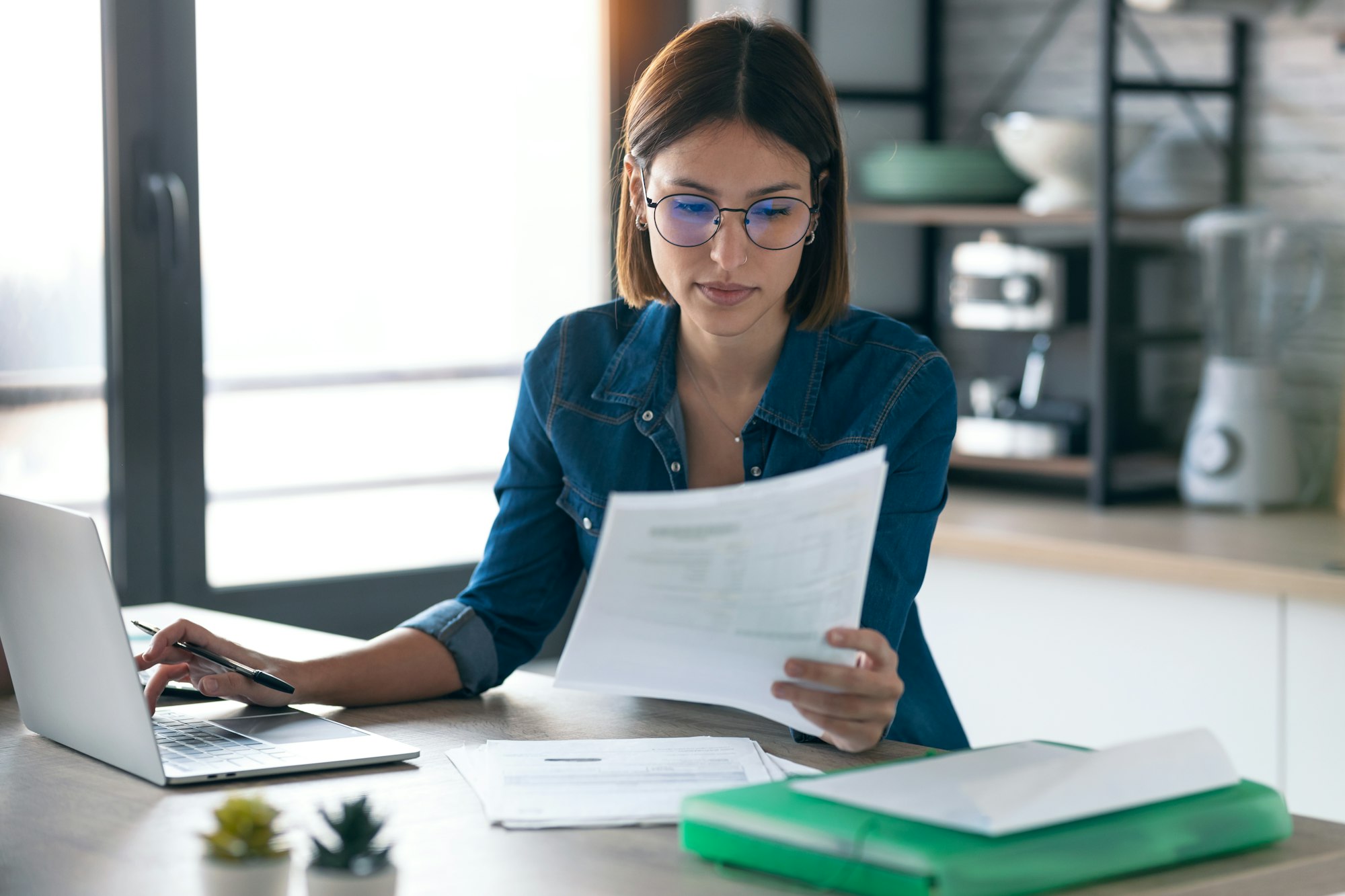 young woman working with computer while consulting some invoices and documents in the kitchen